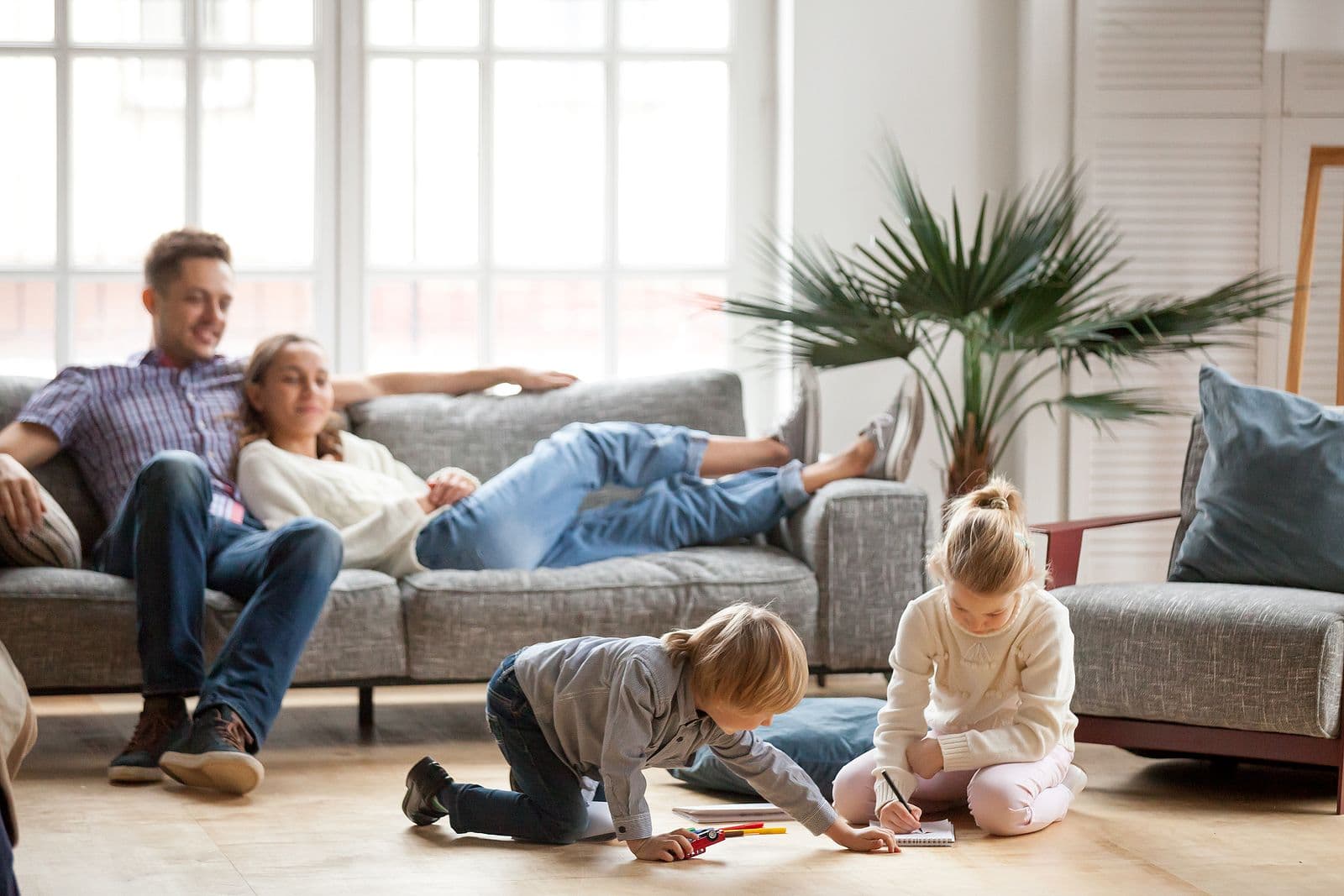 Children Sister And Brother Playing Drawing Together On Floor While Young Parents Relaxing At Home On a Couch.